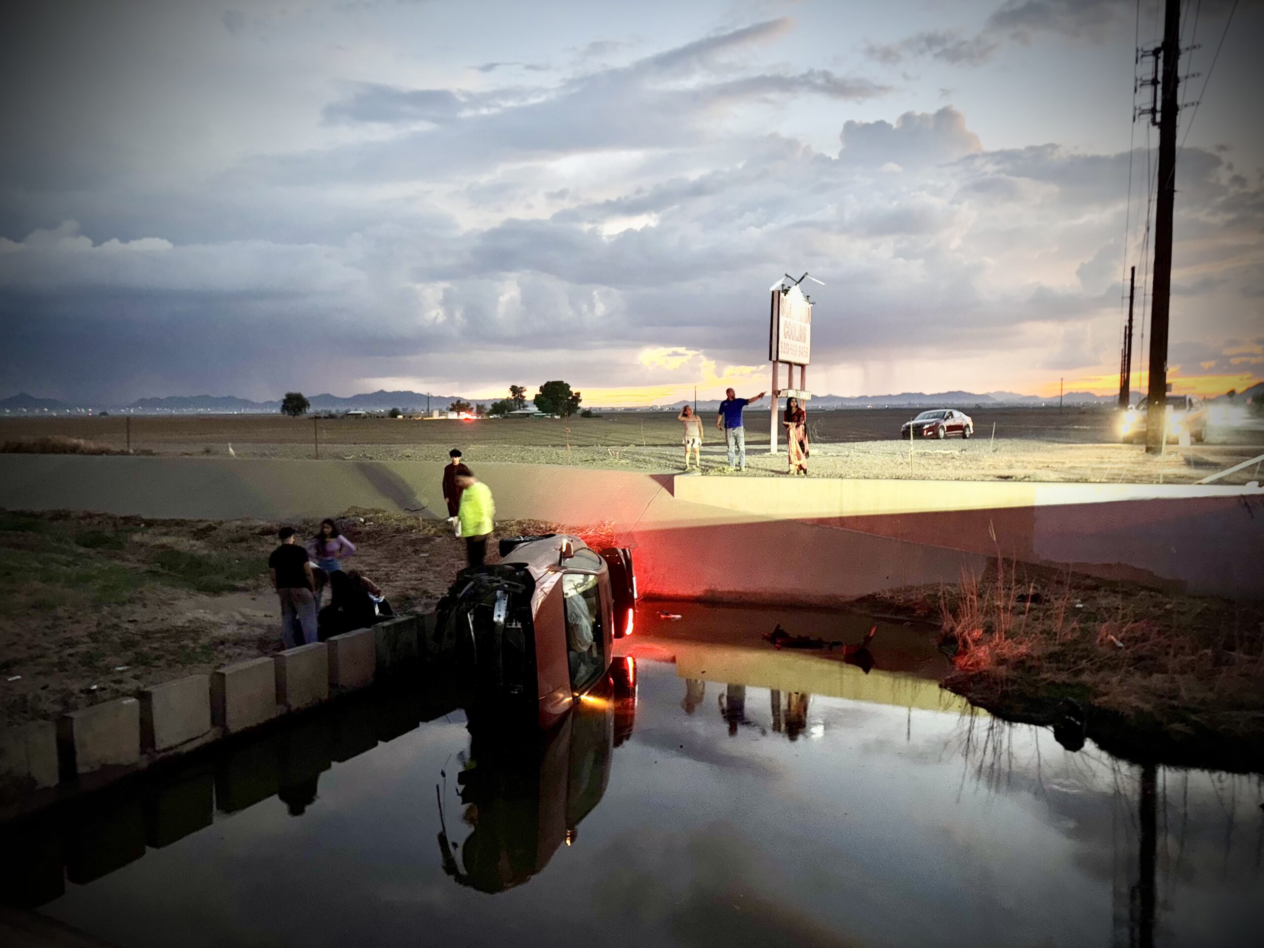 The crashed vehicle rests on its side in muddy water beneath a traffic bridge on Papago Road as crews and bystanders assess the scene. Occupants suffered minor to moderate injuries. [Elias Weiss]